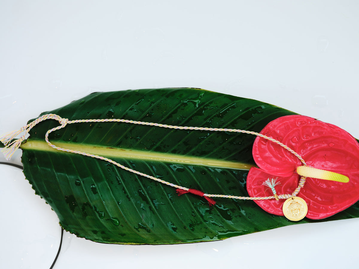 Festival Thread Necklace laid across a tropical leaf with the braided cord and gold pendant shown beside a red anthurium