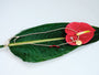 Wide styled view of the Festival Thread Necklace with multicolor woven cord, gold pendant, green leaf, and red anthurium