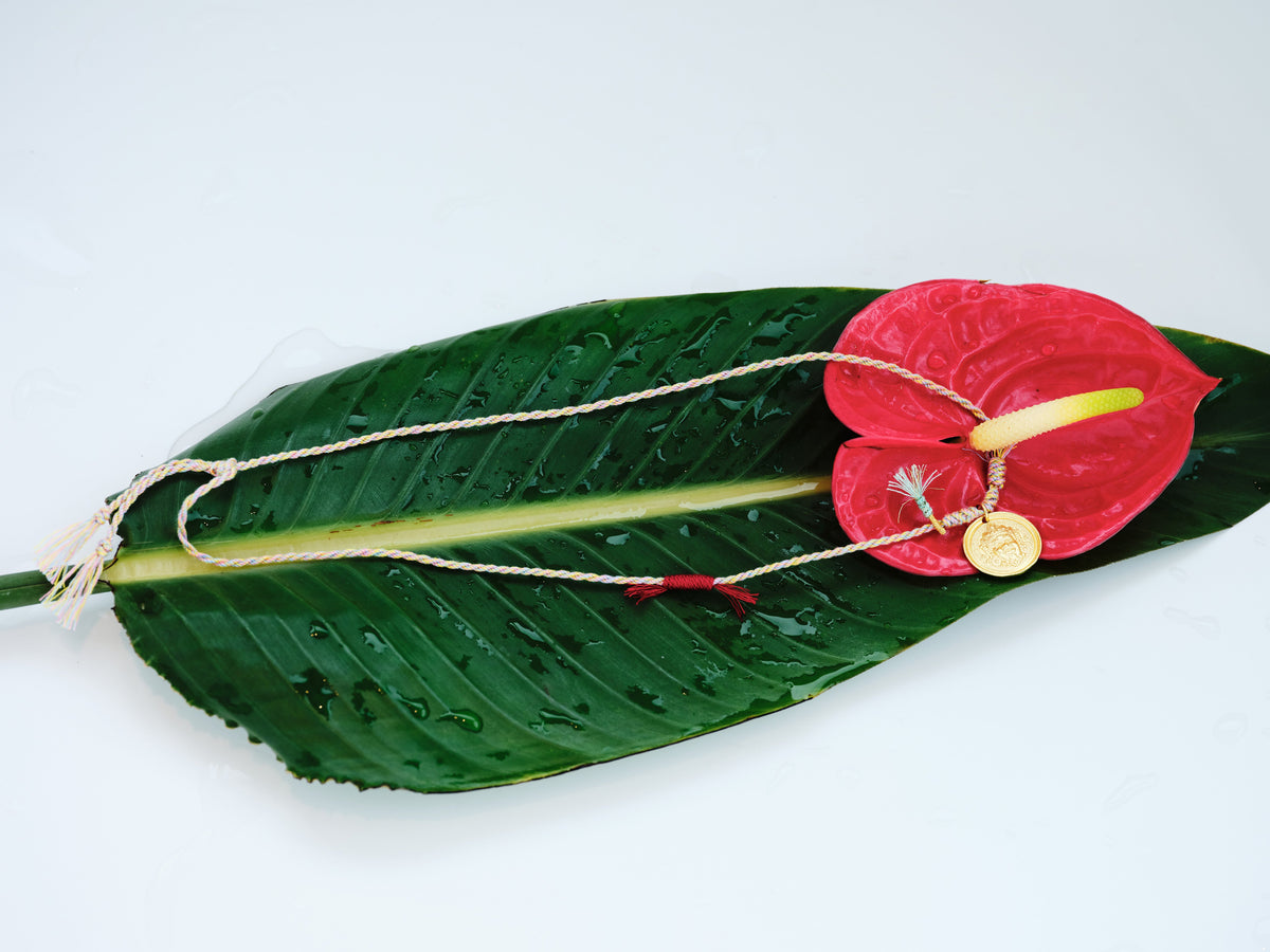 Wide styled view of the Festival Thread Necklace with multicolor woven cord, gold pendant, green leaf, and red anthurium