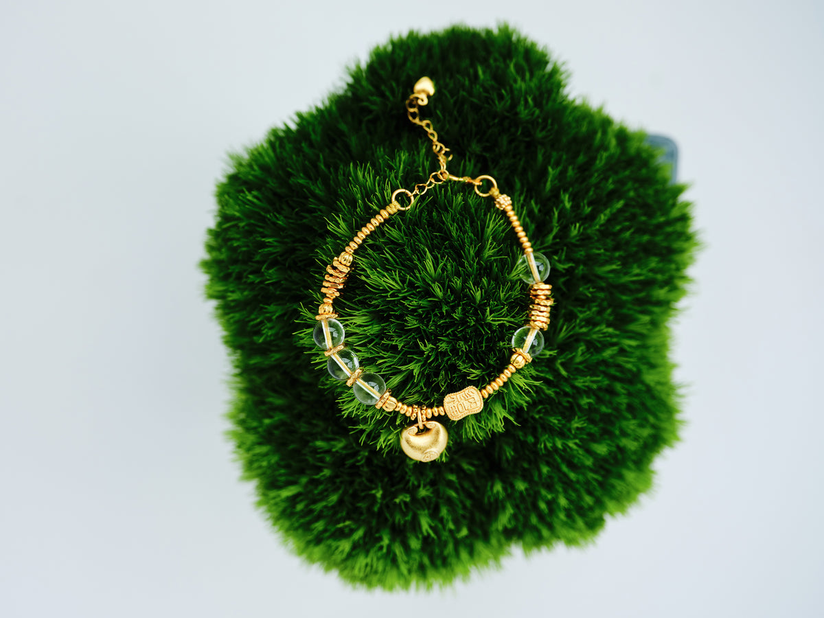 Golden Ingot Current Bracelet with clear crystal beads and a 24K gold-plated copper yuanbao charm on green moss.
